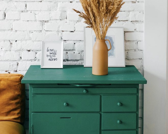 Interior scene with a green dresser against a white brick wall. On the dresser, there is a tan ceramic vase containing dried wheat stalks, flanked by two framed pictures, one reading 'love will keep us alive'.